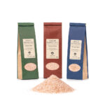 Drei 250g-Beutel mit fein gemahlenem, rosa Kristallsalz in grüner, roter und blauer Verpackung, davor eine kleine Menge des losen Salzes.