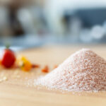 Ein Haufen feines rosa Kristallsalz liegt auf einem Holzbrett, mit bunten Tomaten im Hintergrund.