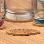 Ein runder Kork-Untersetzer mit eingravierter Blume des Lebens liegt auf einem Holztisch, während im unscharfen Hintergrund Wassergläser auf weiteren Untersetzern stehen.