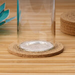 Eine Glaskaraffe steht auf einem runden Korkuntersetzer, der mit dem Symbol der Blume des Lebens graviert ist und auf einem Holztisch liegt.