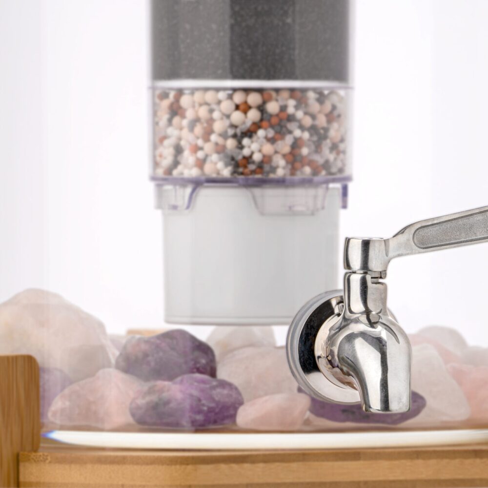 Nahaufnahme eines Edelstahl-Wasserhahns an einem Glas-Wasserspender, im Hintergrund eine mehrstufige Filterkartusche und am Boden Edelsteine.