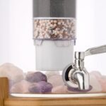 Nahaufnahme eines Edelstahl-Wasserhahns an einem Glas-Wasserspender, im Hintergrund eine mehrstufige Filterkartusche und am Boden Edelsteine.