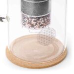 Detailaufnahme einer Wasserfilter-Glaskaraffe mit Blume des Lebens-Gravur und sichtbarer Filterkartusche auf einem Korksockel.