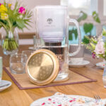 Eine stilvolle Glas-Wasserfilterkanne von Lotus Vita steht auf einem gedeckten Holztisch, davor liegt der goldene Bambusdeckel mit einer Gravur der Lebensblume.