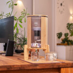 Ein Glas-Wasserfilter mit Bambusgehäuse steht auf einem hölzernen Schreibtisch, umgeben von Zimmerpflanzen.