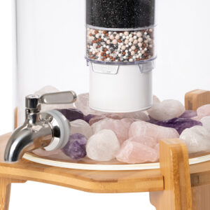 Nahaufnahme eines Glas-Wasserfilters mit Edelstahl-Zapfhahn, einer mehrstufigen Filterkartusche und einer Schicht aus Rosenquarz-, Amethyst- und Bergkristall-Edelsteinen, platziert auf einem Bambusgestell.